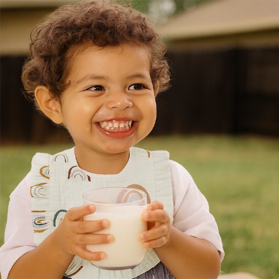 Enfant souriant portant un bavoir bébé coton brodé, tenant un verre de lait dans un jardin.