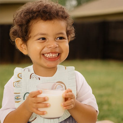 Enfant souriant portant un bavoir bébé coton brodé, tenant un verre de lait dans un jardin.