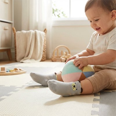 Un bébé heureux portant des chaussettes bebe grises, jouant avec une balle colorée sur un tapis.