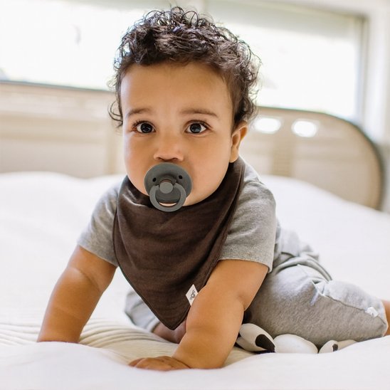 Doudou bebe : un petit garçon avec un bavoir brun et une tétine, jouant sur un lit.